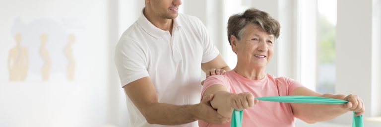 Smiling senior woman stretching with green elastic tape during individual exercises with physiotherapist