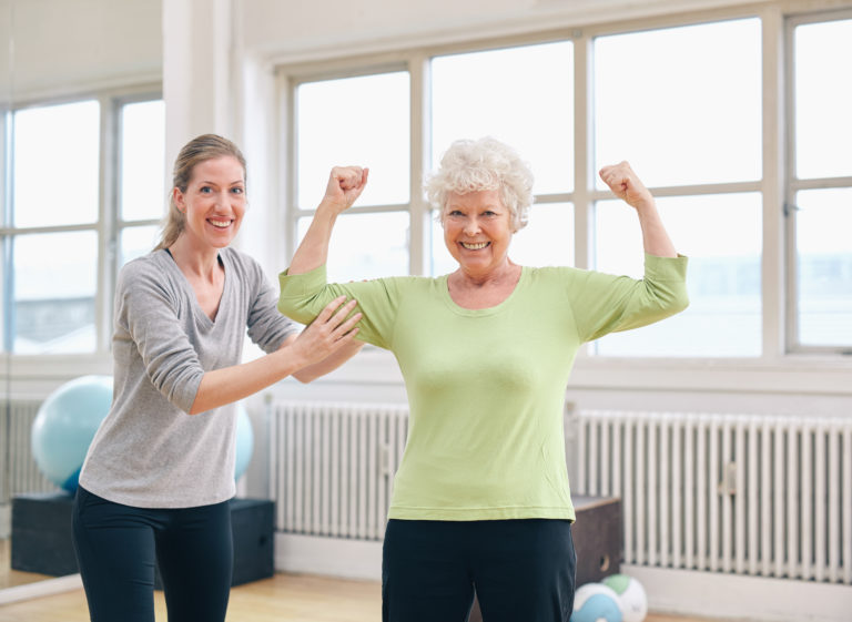 Portrait of a fit old woman flexing her arms and showing her muscles with personal trainer at gym