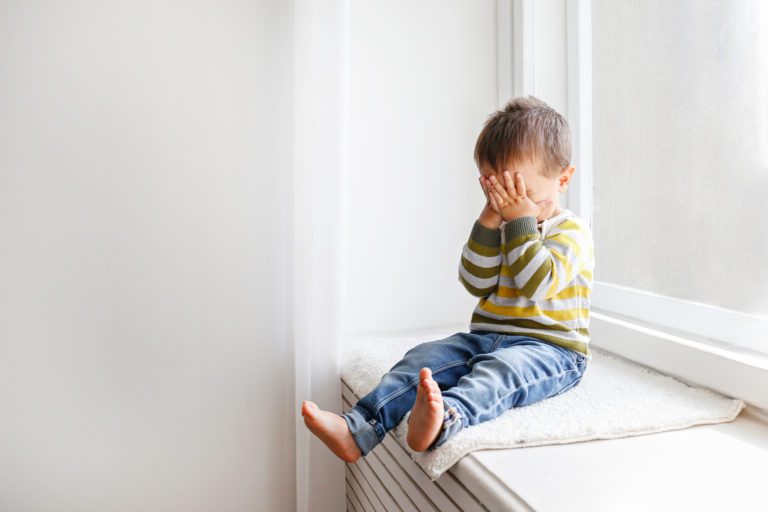 Portrait of adorable little boy sitting on the windowsill and crying. Upset child covering his face at home. Barefoot kid hiding behind palms of his hands. Close up, copy space, background.